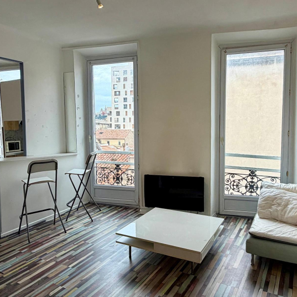 Intérieur d'un petit salon lumineux avec un sol en bois multicolore, deux fenêtres et un mobilier minimaliste.