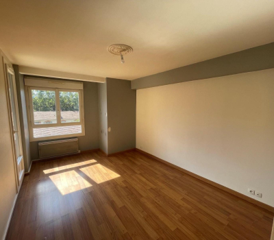 Une pièce vide avec un parquet en bois, des murs blancs et gris, et une fenêtre laissant entrer la lumière du soleil.