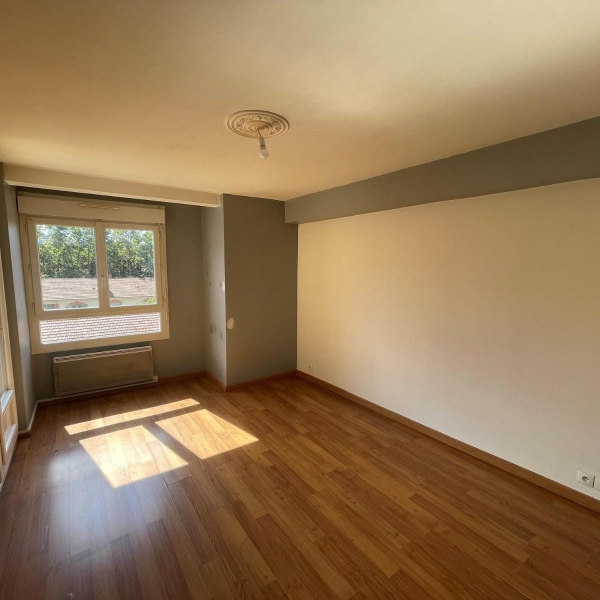 Une pièce vide avec un parquet en bois, des murs blancs et gris, et une fenêtre laissant entrer la lumière du soleil.