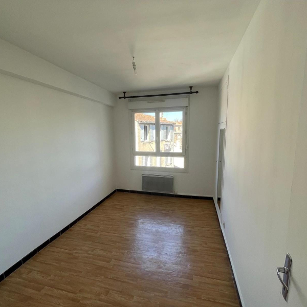 Une pièce vide avec des murs blancs, un sol en parquet et une fenêtre donnant sur d'autres bâtiments.