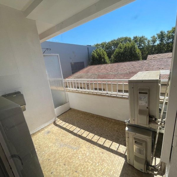 Vue d'un balcon carrelé avec un climatiseur extérieur, surplombant des toits de tuiles et des arbres sous un ciel bleu.