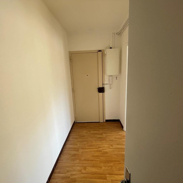 Un couloir étroit avec des murs blancs et un sol en parquet, menant à une porte beige.