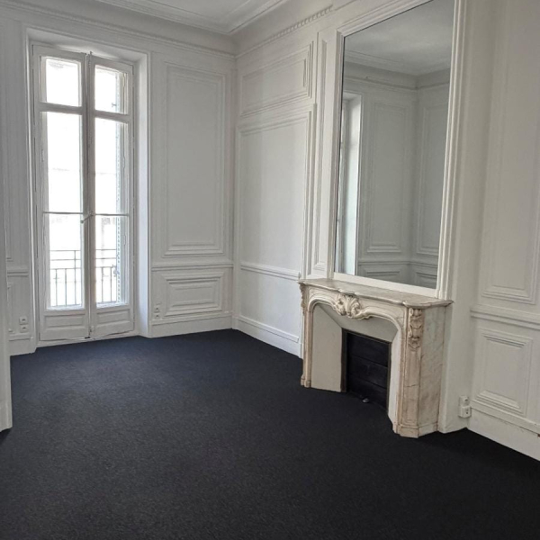 Une pièce vide aux murs blancs avec moulures, un tapis noir, une cheminée en marbre et un grand miroir.