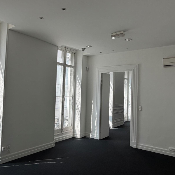 Intérieur d'une pièce vide et lumineuse avec des murs blancs, un sol sombre et de grandes fenêtres.