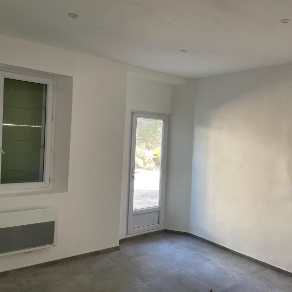 Intérieur d'une pièce vide avec des murs blancs, un carrelage gris, une fenêtre et une porte vitrée.