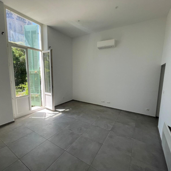 Une pièce vide avec des murs blancs, un sol en carrelage gris et une grande fenêtre ouverte.