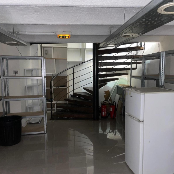 Un sous-sol encombré avec un escalier en colimaçon, des étagères métalliques et un petit réfrigérateur blanc.