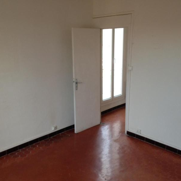 Intérieur d'une pièce vide avec des murs blancs, un sol en terre cuite et une porte ouverte menant à une fenêtre.